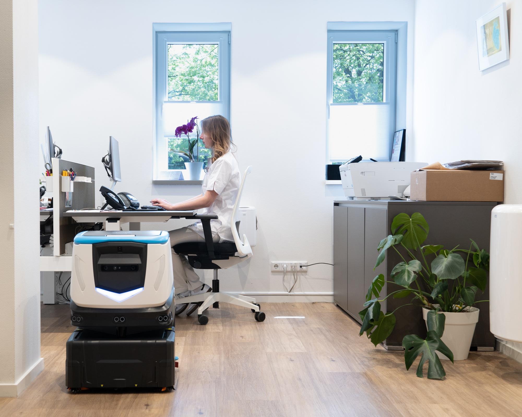 Cobi 18 an autonomous scrubber by ICE Cobotics cleaning healthcare office while healthcare employee works on computer in background