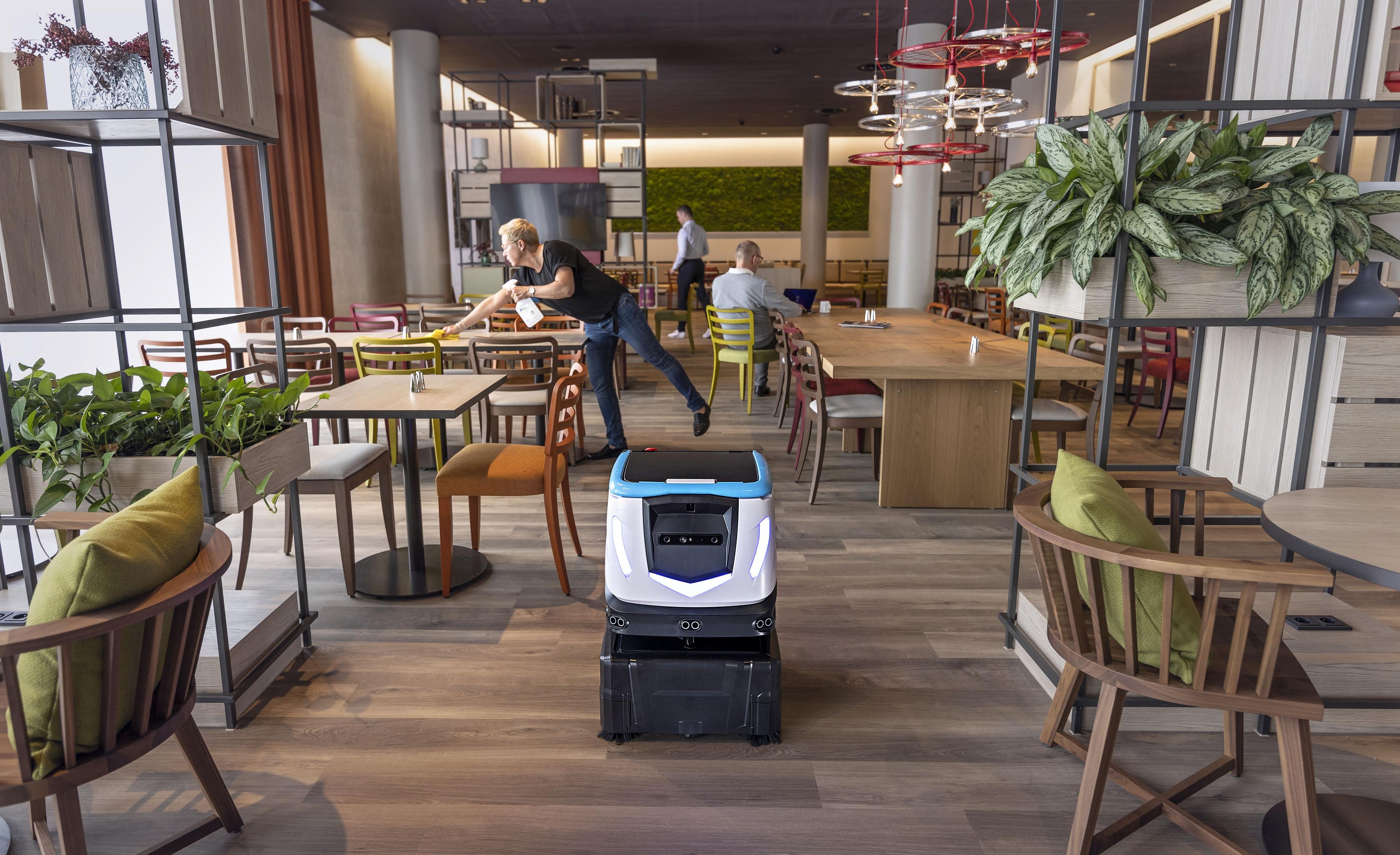 ICE Cobotics Cobi 18 cleaning floors in hotel restaurant while workers clean tables and guests eat breakfast