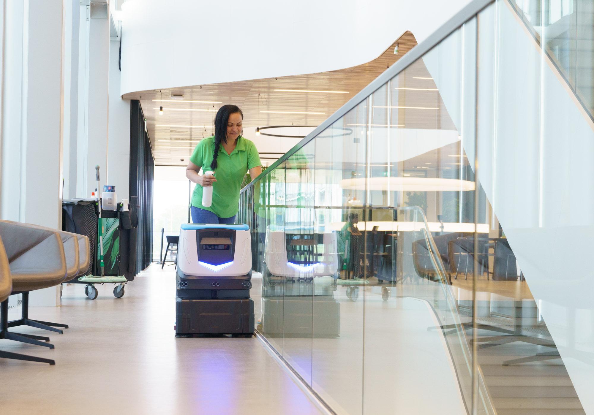 ICE Cobotics Cobi 18, robotic floor scrubber, cleans commercial office building next to cleaning staff member wiping down surfaces