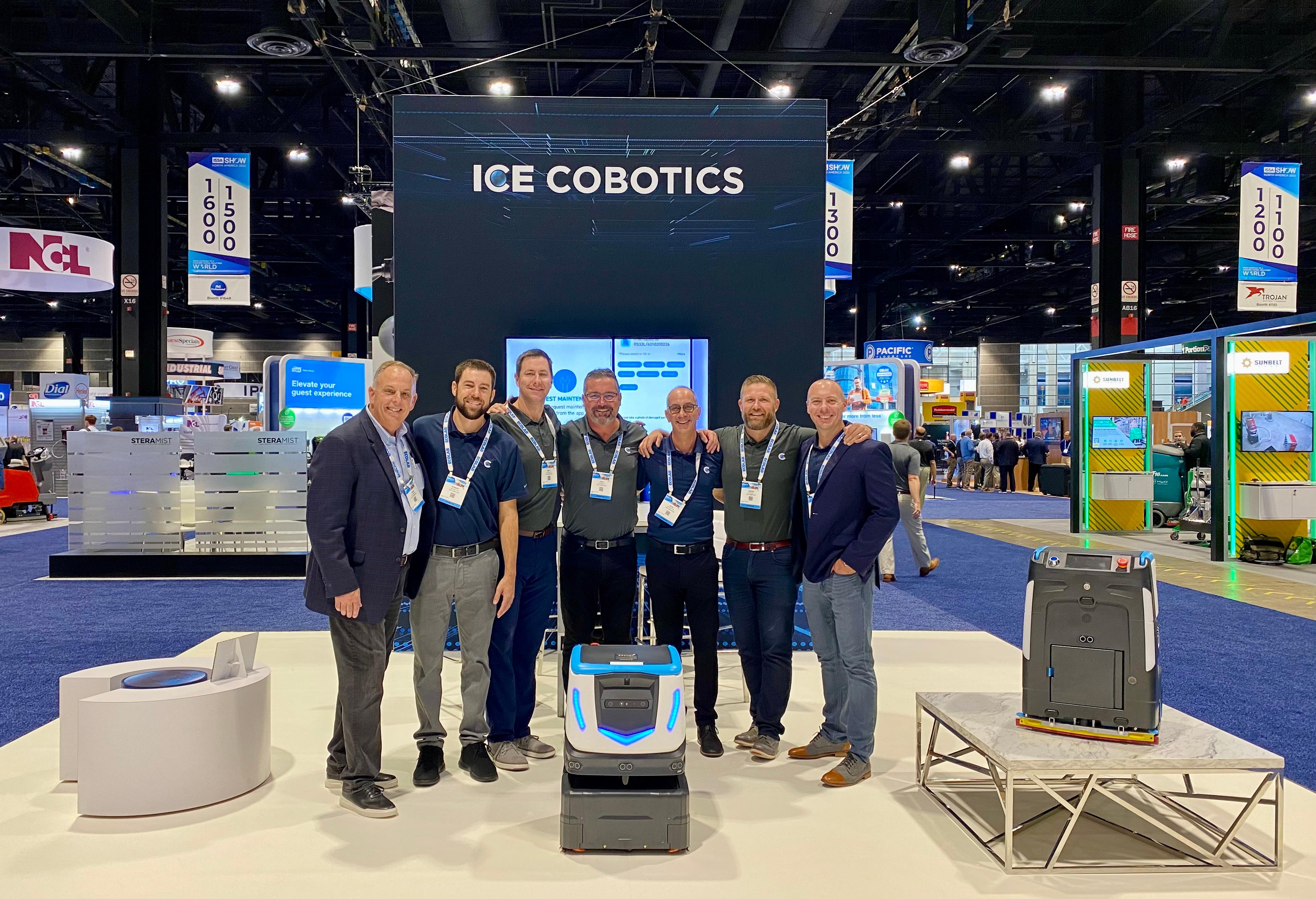 ICE Cobotics ISSA 2022 team memberst standing in trade show booth in front of large video wall and next to Cobi 18, robotic floor scrubber